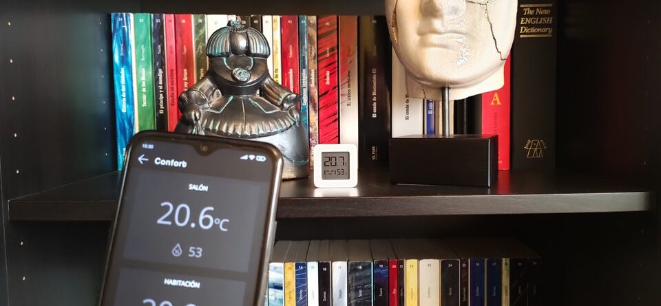 Móvil en primer plano con la pantalla de temperaturas de la app Qvadis y al fondo un termómetro Xiaomi Mijia en una estantería de un salón